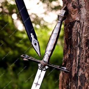 May include: A detailed image of a medieval sword with a silver blade and ornate hilt. The sword is partially sheathed in a black scabbard. The sword is leaning against a tree trunk, with a blurred green background.