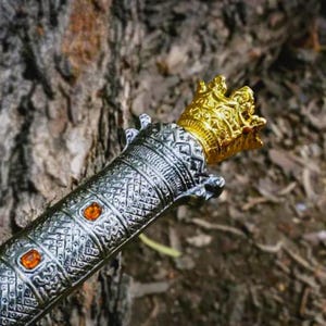 Puede incluir: Primer plano de una empu&ntilde;adura de espada decorativa con una corona dorada y un mango plateado. El mango presenta un intrincado patr&oacute;n y gemas naranjas. La espada es probablemente un art&iacute;culo decorativo o un accesorio.