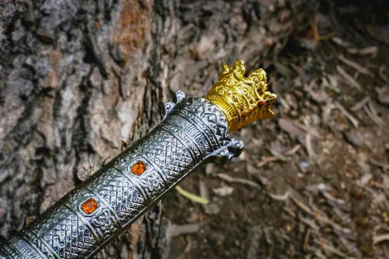 Puede incluir: Primer plano de una empu&ntilde;adura de espada decorativa con una corona dorada y un mango plateado. El mango presenta un intrincado patr&oacute;n y gemas naranjas. La espada es probablemente un art&iacute;culo decorativo o un accesorio.