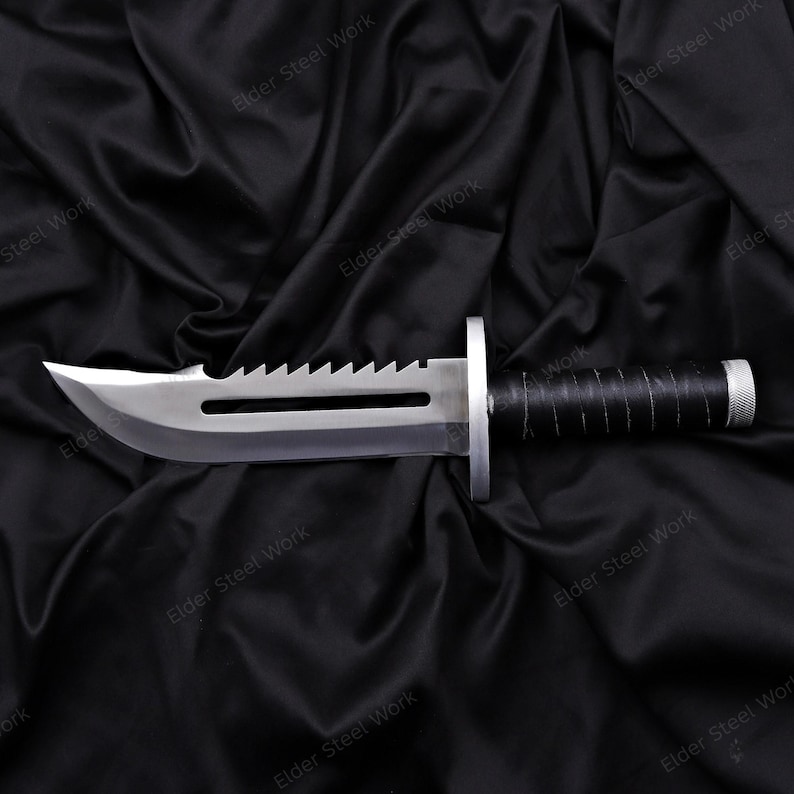 May include: A stainless steel survival knife with a serrated spine and a black wrapped handle. The blade has a sharp edge and a fuller. The knife is displayed on a black satin background.