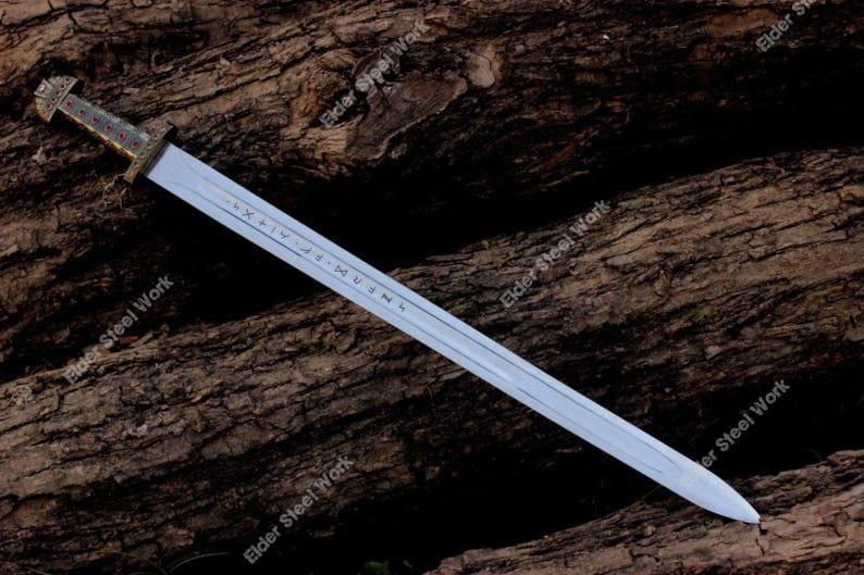May include: A long, silver sword with a decorative handle featuring red and gold accents. The blade has etched markings and is displayed against a textured, brown wooden surface. The words "Elder Steel Work" are visible.