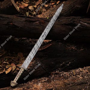 May include: A long, ornate sword with a patterned blade and a detailed hilt. The blade has a textured, wavy design. The hilt features red accents and a decorative pommel. The sword rests on a dark wooden surface.