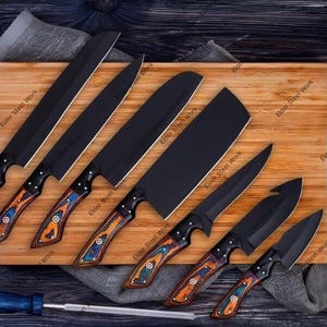 Op de afbeelding: Een verzameling van zeven zwarte keukenmessen met houten handvatten met een kleurrijk patroon. De messen liggen op een houten snijplank, met een blauw handvat gereedschap onderaan. De messen variëren in grootte en vorm.
