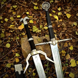May include: Two silver and black swords with detailed designs, including a wolf head and a circular emblem. The blades are displayed on autumn leaves, highlighting the craftsmanship and detail of the weapons.