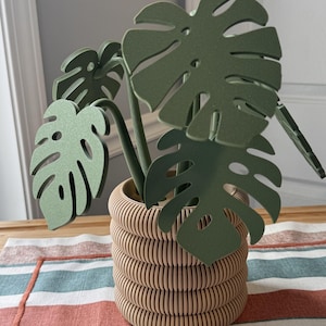 Pode incluir: Uma escultura decorativa de planta com folhas de monstera planas, recortadas e de cor verde-escura com perfurações características, sustentadas por caules que emergem de um vaso cilíndrico de cor bege com textura canelada.