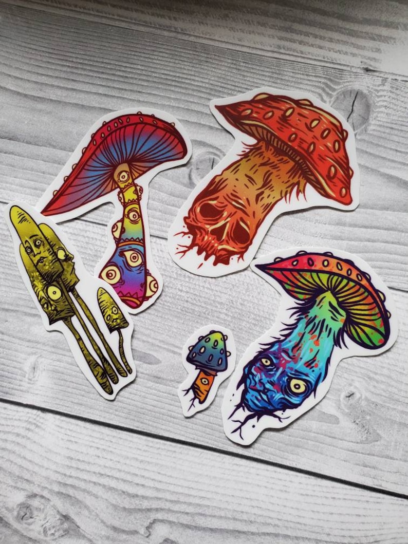 May include: A collection of colorful mushroom stickers with unique designs. The stickers feature various styles, including psychedelic and skull-like illustrations, with vibrant hues of red, blue, green, and yellow. The stickers are on a light gray wooden surface.