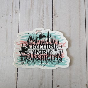 May include: A white sticker with a stylized forest scene and the text "CRYPTIDS FOR TRANS RIGHTS" in black. The design includes a flag with the transgender pride colors, silhouettes of creatures, and a pink and blue sky.