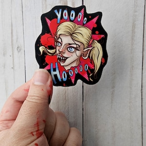 May include: A die-cut sticker featuring a blonde character with pointed ears and sharp teeth, set against a black background with a red comic-style starburst. The text YOOO HOOOO is printed in blue with a white outline above and below the character.