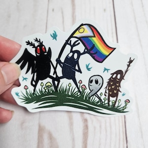 May include: A sticker featuring four cartoon characters, including a black moth, a deer, a ghost, and a brown creature with a stick. The deer is holding a rainbow pride flag with a yellow circle in the top left corner. The characters are standing on a green grassy hill with pink and yellow mushrooms.