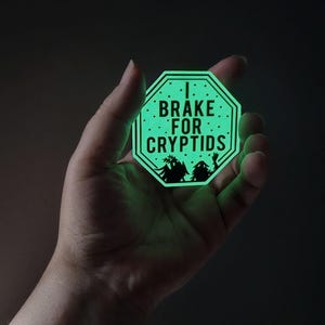 May include: A glowing green octagon sticker with the text "I Brake For Cryptids" in white. The sticker features black silhouettes of cryptids. The sticker is held in a hand.
