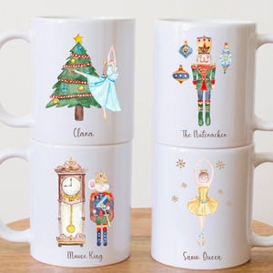 May include: Four white ceramic mugs with festive illustrations. Designs include a Christmas tree with a ballerina, a Nutcracker, a clock with a mouse king, and a ballerina labelled "Snow Queen". Each mug has a white handle and is stacked on a wooden surface.