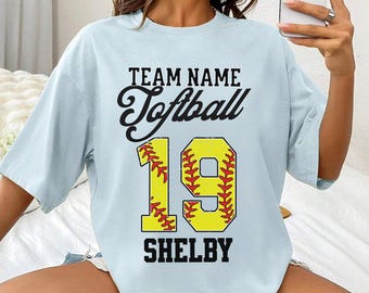 May include: Pale blue t-shirt featuring the text "TEAM NAME Softball 19 SHELBY" in black and yellow. The number 19 is designed to resemble a softball. The shirt has a relaxed, oversized style.