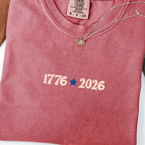 May include: A dusty rose t-shirt with the numbers "1776" and "2026" in a cream color, separated by a blue star. A gold-colored necklace with a small pendant rests on the shirt. The shirt is folded, and a brown hat is partially visible.