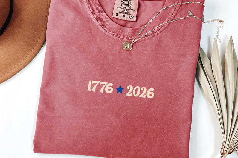May include: A dusty rose t-shirt with the numbers "1776" and "2026" in a cream color, separated by a blue star. A gold-colored necklace with a small pendant rests on the shirt. The shirt is folded, and a brown hat is partially visible.