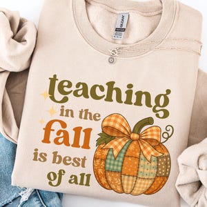 May include: Beige sweatshirt with the text "teaching in the fall is best of all" in green and orange. A patchwork pumpkin graphic is featured below the text. A silver necklace is visible.