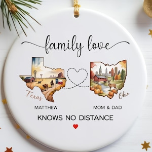 May include: A round white ceramic ornament featuring the text family love and KNOWS NO DISTANCE. It displays a map of Texas with a rural scene and a map of Ohio with a landscape, connected by a dashed heart line. Names Matthew and Mom & Dad are printed below.