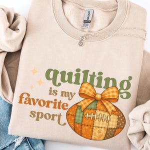 Könnte beinhalten: Beiges Sweatshirt mit dem Text "quilting is my favorite sport" in Orange und Grün. Ein Patchwork-Football mit einer orangefarbenen Schleife ist im Design enthalten. Das Sweatshirt ist gefaltet, eine Jeans ist sichtbar.