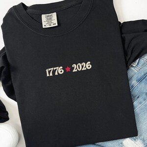 May include: Black long-sleeve shirt with the text "1776 2026" in a cream color with a red star. The shirt is paired with blue jeans and white sneakers. The shirt has a Comfort Colors tag.
