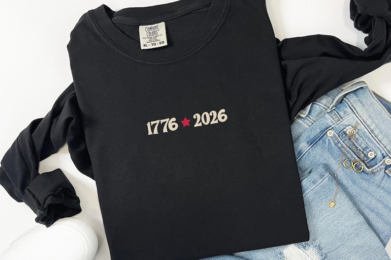 May include: Black long-sleeve shirt with the text "1776 2026" in a cream color with a red star. The shirt is paired with blue jeans and white sneakers. The shirt has a Comfort Colors tag.