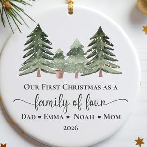 May include: A round white ceramic ornament featuring a watercolor illustration of four green pine trees. The text reads: Our First Christmas as a family of four, Dad, Emma, Noah, Mom, and the year 2026. It hangs from a gold string.