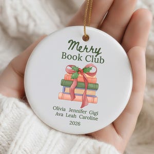 Pode incluir: Um ornamento redondo de cer&acirc;mica branca com o texto verde Merry Book Club sobre uma ilustra&ccedil;&atilde;o de uma pilha de livros atados com uma fita vermelha e azevinho. Abaixo dos livros est&atilde;o os nomes Olivia, Jennifer, Gigi, Ava, Leah, Caroline e 2026.