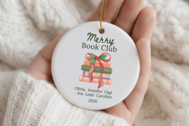 Pode incluir: Um ornamento redondo de cer&acirc;mica branca com o texto verde Merry Book Club sobre uma ilustra&ccedil;&atilde;o de uma pilha de livros atados com uma fita vermelha e azevinho. Abaixo dos livros est&atilde;o os nomes Olivia, Jennifer, Gigi, Ava, Leah, Caroline e 2026.