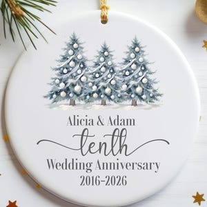 May include: A round white ceramic ornament featuring a watercolor illustration of three blue-toned Christmas trees decorated with white ornaments. The text reads Alicia & Adam, tenth Wedding Anniversary, 2016-2026 in dark gray lettering.