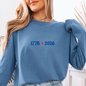 May include: A long-sleeved, blue sweatshirt with the text "1776 2026" in blue and a red star. The sweatshirt is made of a soft material and has a crew neck. The overall theme is patriotic.
