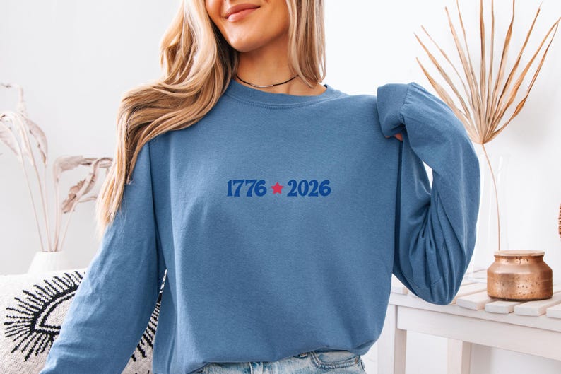 May include: A long-sleeved, blue sweatshirt with the text "1776 2026" in blue and a red star. The sweatshirt is made of a soft material and has a crew neck. The overall theme is patriotic.