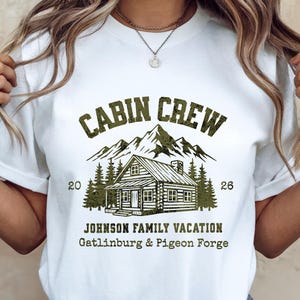 Puede incluir: Camiseta blanca con un gr&aacute;fico verde que dice "CABIN CREW" sobre una ilustraci&oacute;n de caba&ntilde;a, monta&ntilde;as y &aacute;rboles. Debajo del gr&aacute;fico est&aacute; "JOHNSON FAMILY VACATION Gatlinburg & Pigeon Forge". La camiseta tiene los n&uacute;meros "20" y "26".