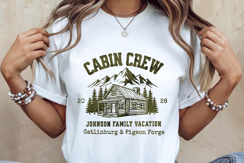 Puede incluir: Camiseta blanca con un gr&aacute;fico verde que dice "CABIN CREW" sobre una ilustraci&oacute;n de caba&ntilde;a, monta&ntilde;as y &aacute;rboles. Debajo del gr&aacute;fico est&aacute; "JOHNSON FAMILY VACATION Gatlinburg & Pigeon Forge". La camiseta tiene los n&uacute;meros "20" y "26".