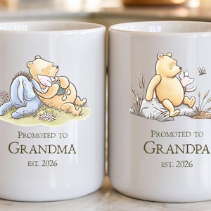 May include: Two white ceramic mugs featuring Winnie the Pooh illustrations. The left mug shows Pooh and Eeyore with the text PROMOTED TO GRANDMA EST. 2026. The right mug shows Pooh and Piglet with the text PROMOTED TO GRANDPA EST. 2026.