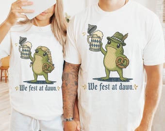 May include: A white t-shirt featuring a graphic of a green frog holding a beer stein and a pretzel. The frog on the left wears a blonde braided wig, while the frog on the right wears a green hat with a feather. The text "We fest at dawn." is printed below.
