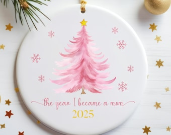 May include: A white ceramic Christmas ornament with a pink watercolor Christmas tree design, snowflakes, and the text "the year I became a mom 2025". The ornament has a gold hanging loop and is surrounded by gold star and ball decorations.