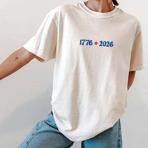 May include: Cream-colored t-shirt with the blue text "1776 * 2026" centered on the chest. The design includes a blue star between the numbers. The shirt is paired with blue jeans.