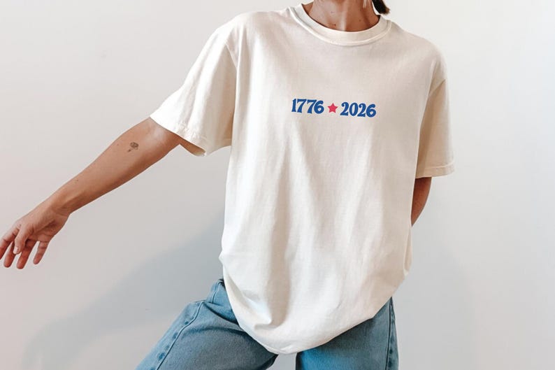 May include: Cream-colored t-shirt with the blue text "1776 * 2026" centered on the chest. The design includes a blue star between the numbers. The shirt is paired with blue jeans.
