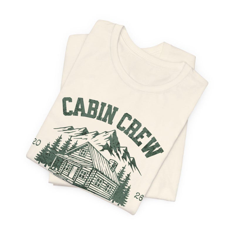 Puede incluir: Camiseta color crema con un gr&aacute;fico verde que dice "CABIN CREW" sobre una ilustraci&oacute;n de caba&ntilde;a, monta&ntilde;as y &aacute;rboles. La camiseta est&aacute; doblada y tiene los n&uacute;meros "20" y "26" a los lados del gr&aacute;fico. La camiseta es de material suave.