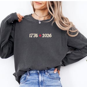 May include: Dark gray long-sleeve sweatshirt with the text "1776" and "2026" separated by a red star. The sweatshirt is made of a soft material and is suitable for casual wear. The model is wearing blue jeans.