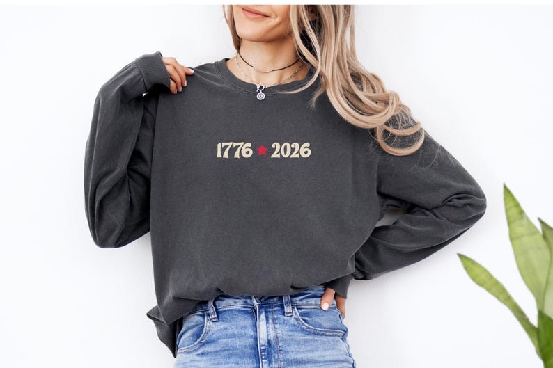 May include: Dark gray long-sleeve sweatshirt with the text "1776" and "2026" separated by a red star. The sweatshirt is made of a soft material and is suitable for casual wear. The model is wearing blue jeans.