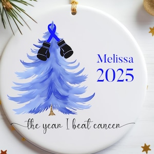 Puede incluir: Adorno navideño redondo blanco con un diseño de árbol de Navidad azul acuarela. El adorno tiene el texto "Melissa 2025" y "the year I beat cancer". El árbol está decorado con una cinta azul y guantes de boxeo negros.