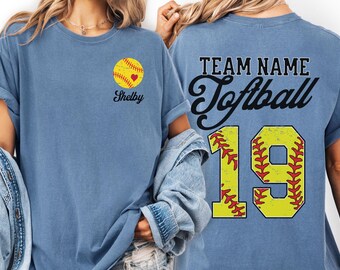 May include: A blue short-sleeved t-shirt with a softball design. The front has a yellow softball with a red heart and the name "Shelby". The back shows "Team Name Softball" and the number "19".