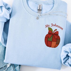 May include: A light blue sweatshirt with the name "Ms. Johnson" and a patchwork apple design. The apple features a red bow and various plaid patterns. A silver necklace is draped across the neckline. The sweatshirt is paired with blue jeans.