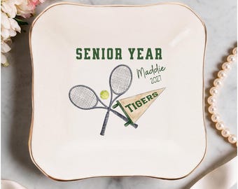 Pode incluir: Um prato quadrado de cerâmica branca com borda dourada e um design de ténis. Inclui duas raquetes cruzadas, uma bola de ténis, uma flâmula com a palavra TIGERS e o texto em verde SENIOR YEAR, Maddie e 2027 impresso no centro.