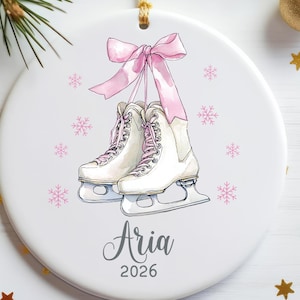 May include: A round white ceramic ornament featuring a watercolor illustration of white ice skates tied together with a pink ribbon. The design includes small pink snowflakes and the name Aria printed in grey script above the year 2026.