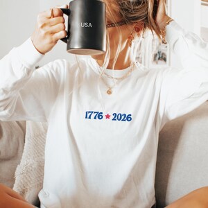 May include: A white long-sleeve shirt with the text "1776 2026" in blue. A black mug with "USA" printed on it is held up. The person is sitting on a couch.