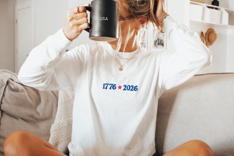 May include: A white long-sleeve shirt with the text "1776 2026" in blue. A black mug with "USA" printed on it is held up. The person is sitting on a couch.