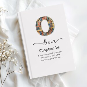 May include: A white hardcover book featuring a large letter O filled with a colorful bookshelf illustration. Below the letter, the name Olivia is printed in script, followed by the text Chapter 14 and A new chapter of laughter, adventure, and a few surprise plot twists.