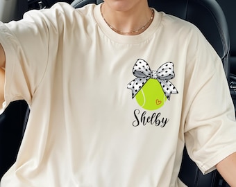 May include: Cream-colored t-shirt featuring a tennis ball design with a polka-dot bow and the name "Shelby" in script. The tennis ball is green with a small heart. The shirt has short sleeves.