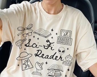 May include: Cream-colored t-shirt with a black graphic design featuring the text "Sci-Fi Reader" and illustrations of books, a ray gun, a robot, an alien, and a satellite dish. The shirt has short sleeves.
