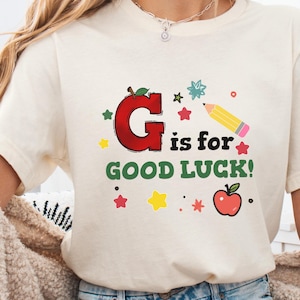 May include: Cream-colored t-shirt with the phrase "G is for GOOD LUCK!" in a playful, colorful design. Features a large red letter "G" shaped like an apple, stars, a pencil, and an additional apple illustration.